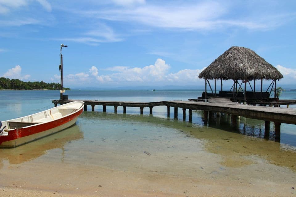 Un bateau rouge et blanc est amarré à une jetée en bois avec une cabane au toit de chaume au-dessus d'une eau tropicale claire sous un ciel bleu.