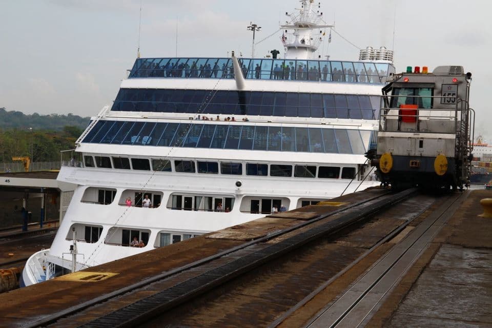 Un train de mules sur rails tire un grand navire de croisière à travers une écluse de canal tandis que les passagers regardent depuis les ponts supérieurs.