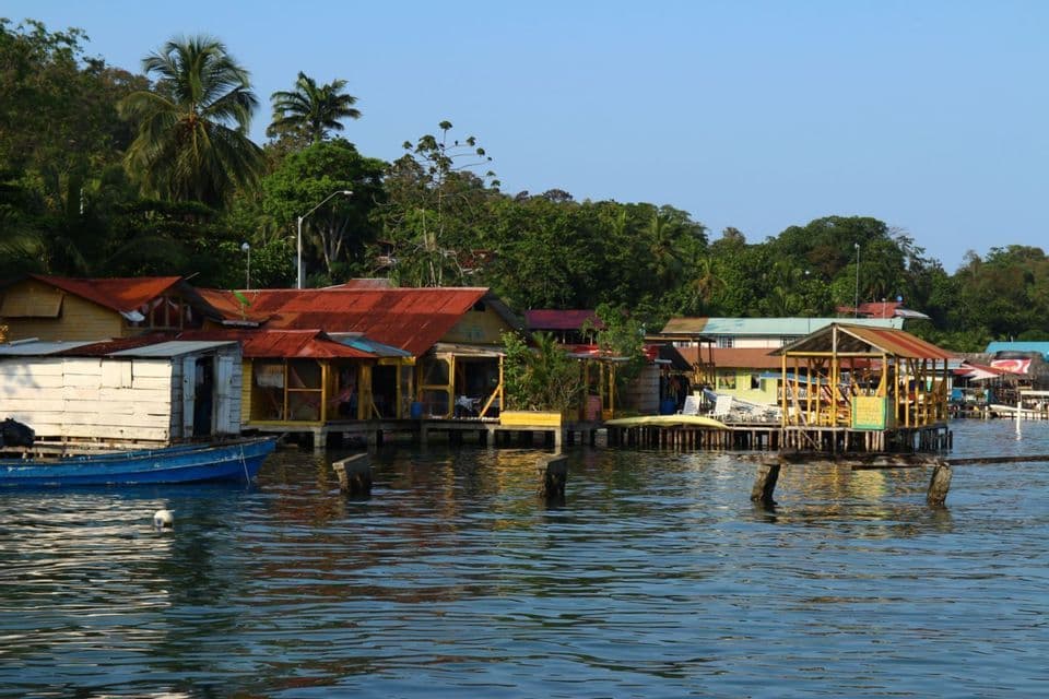 Un village au bord de l'eau avec des maisons colorées sur pilotis, à l'orée d'une forêt luxuriante, avec un petit bateau bleu amarré à proximité.