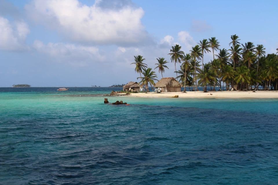 Vue de l'eau d'une île tropicale avec des paillotes et des palmiers sur une plage de sable blanc sous un ciel nuageux.