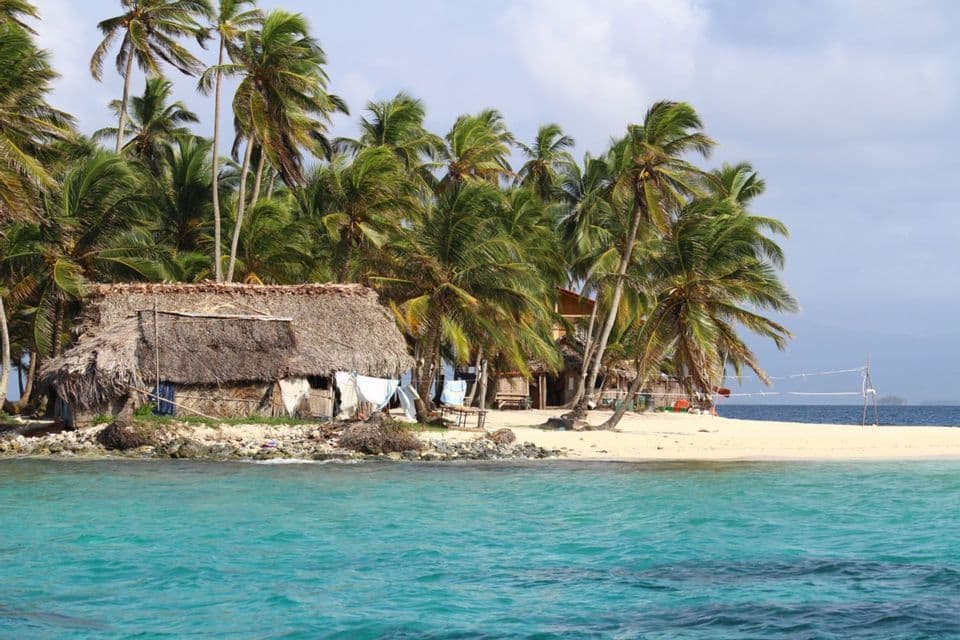 Une cabane au toit de chaume se dresse sur une petite île de sable entourée de palmiers et d'eau turquoise claire.