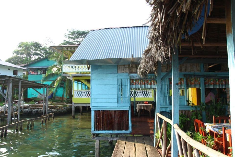 Maisons colorées en bois sur pilotis d'un village construit sur l'eau, avec un restaurant sur une passerelle en bois au premier plan.