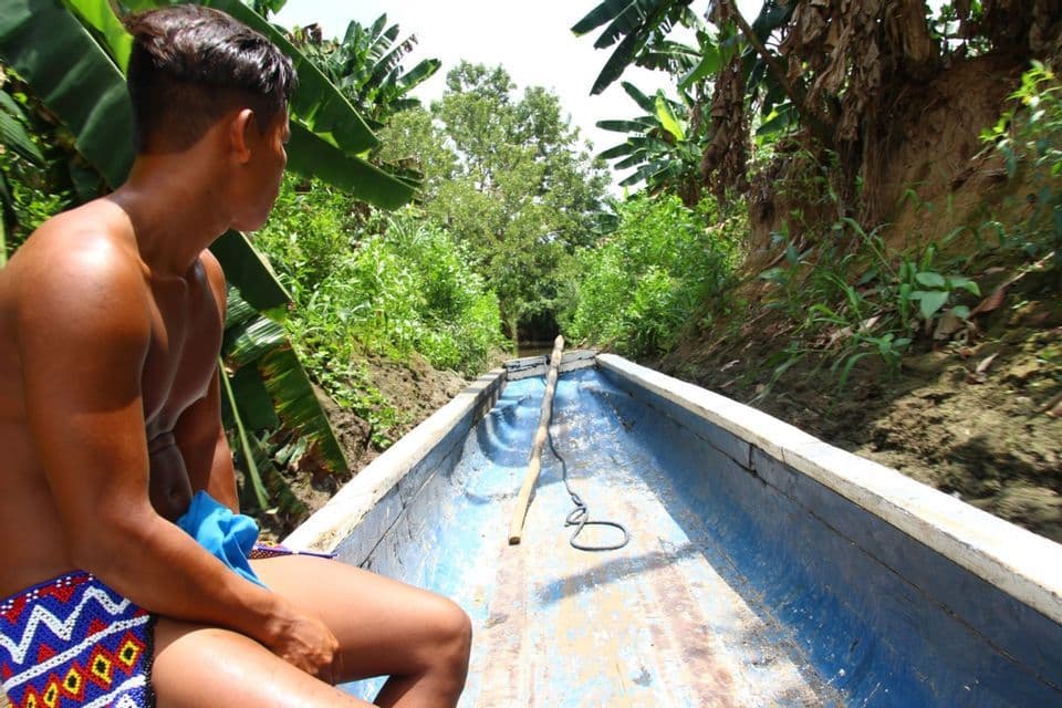 Un homme en short à motifs est assis dans un long bateau bleu, naviguant sur un étroit canal entouré de végétation tropicale.