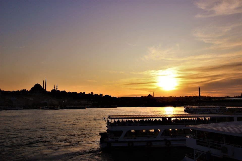 Barcos turísticos con pasajeros navegan sobre el agua al atardecer, con un perfil urbano de la ciudad y mezquitas silueteadas al fondo.