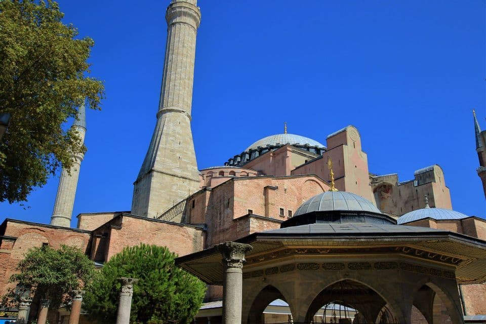 Vista exterior de una gran mezquita con cúpulas y un minarete bajo un cielo azul sin nubes, con una fuente en primer plano.