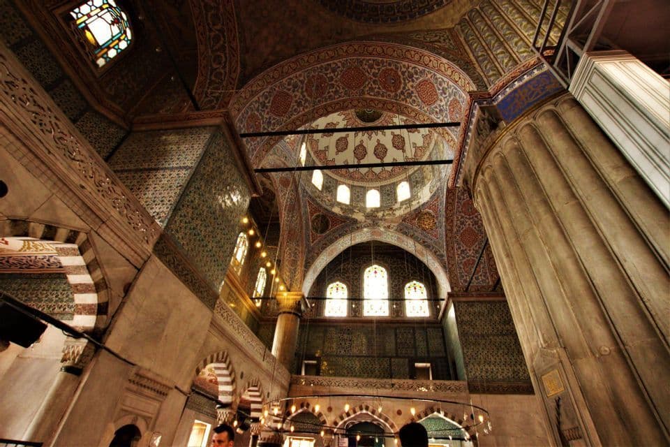 Vista en ángulo bajo del ornamentado interior de una mezquita con cúpulas decoradas, pilares macizos, ventanas arqueadas y azulejos detallados.