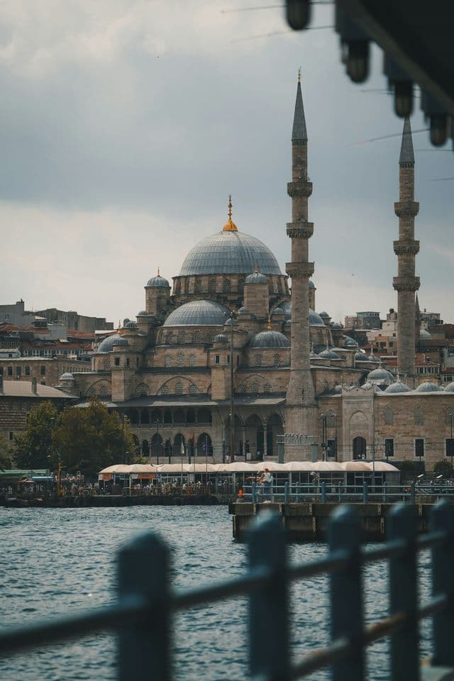 Una gran mezquita con múltiples cúpulas y minaretes se alza a orillas del agua bajo un cielo nublado, vista desde el otro lado del agua.