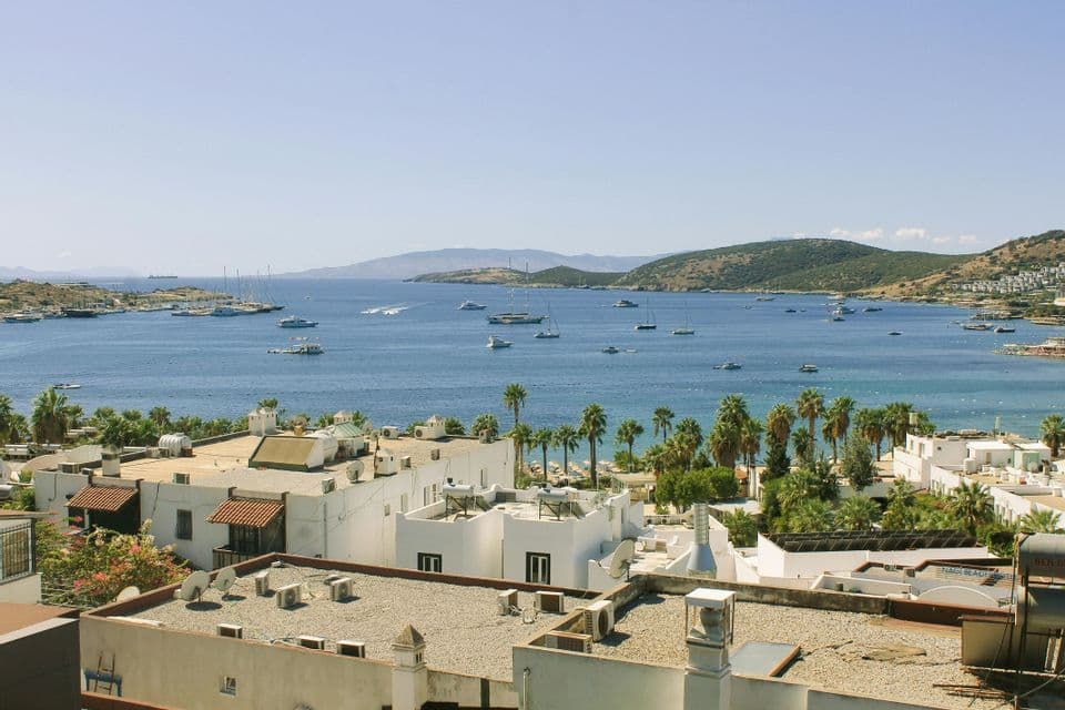 Vista dall'alto di candidi edifici costieri che si affacciano su una placida baia blu, costellata di numerosi yacht e imbarcazioni, sotto un cielo sereno.