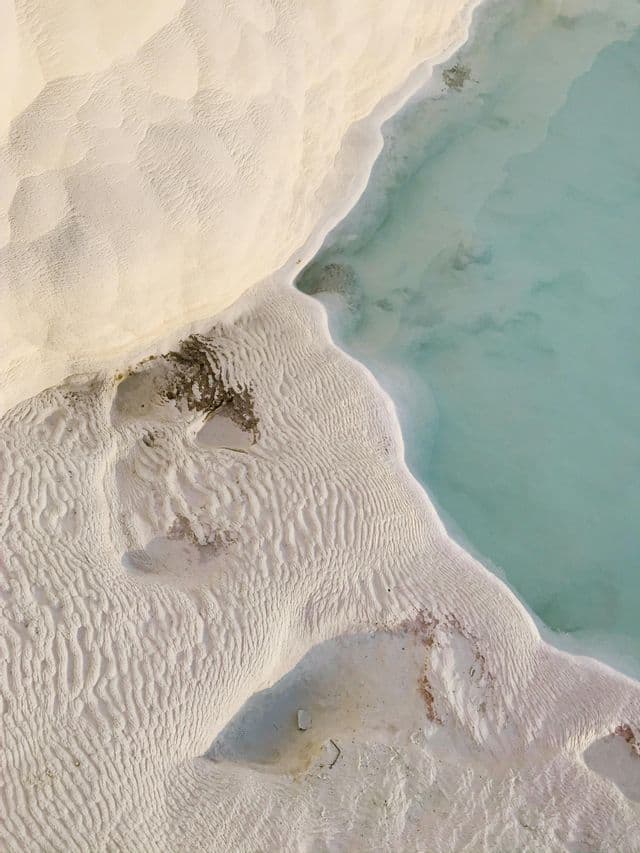 Una vista aerea di depositi minerali bianchi e texturizzati che costeggiano uno specchio d'acqua calma e turchese.