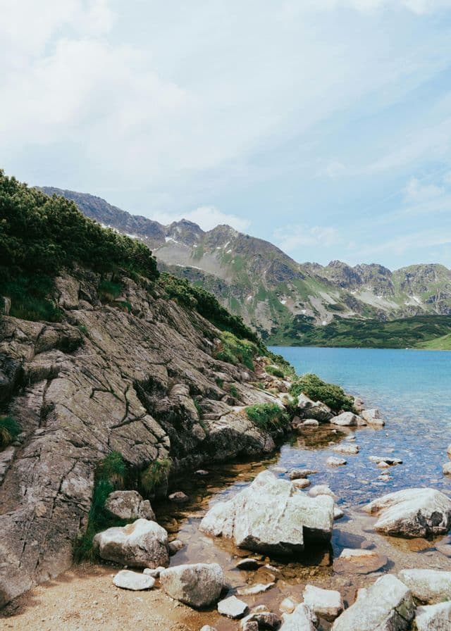 Ein felsiges Ufer trifft auf das klare Wasser eines blauen Alpensees, mit grünen Bergen im Hintergrund unter bewölktem Himmel.