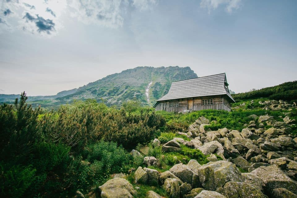 Eine rustikale Holzhütte liegt an einem grünen, felsigen Berghang mit einem großen Berggipfel im Hintergrund unter einem teils bewölkten Himmel.