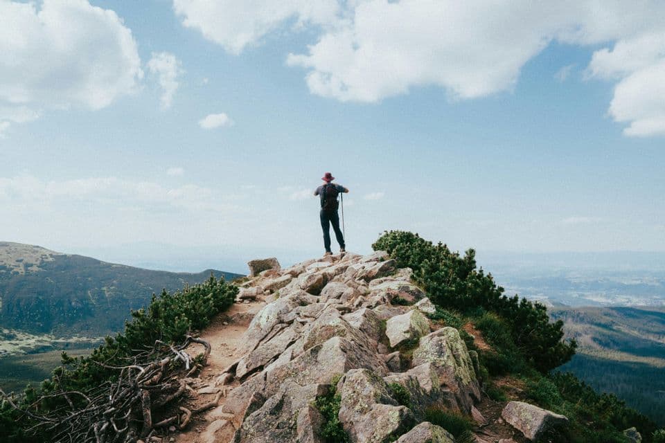 Ein Wanderer mit Rucksack und Trekkingstock steht auf einem felsigen Berggipfel und blickt auf eine weite Landschaft unter blauem Himmel mit Wolken.