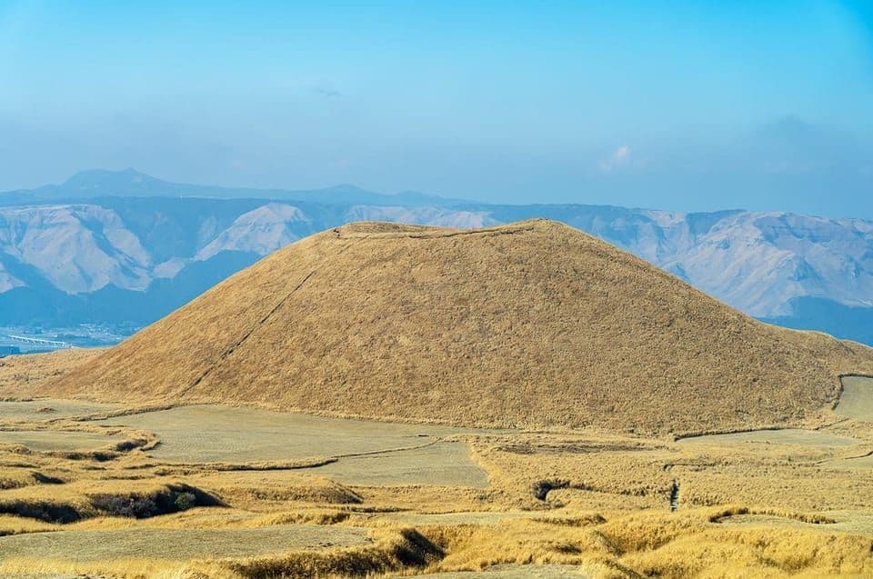 Una grande collina rotonda coperta di erba dorata si erge in un vasto paesaggio con montagne distanti sotto un cielo blu.