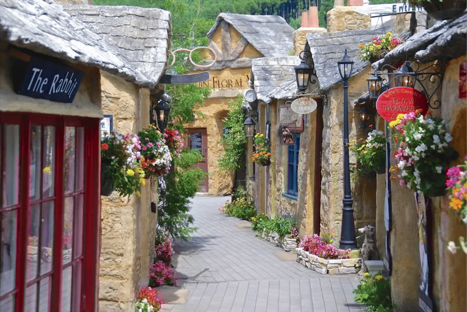 Un vicolo stretto e lastricato si snoda tra edifici in pietra con tetti in ardesia, decorato con fiori colorati, insegne e lampioni.
