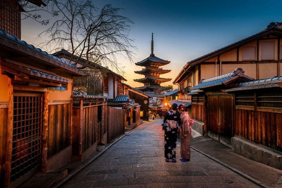 Due donne in kimono tradizionali si trovano su una strada acciottolata in pendenza, affiancata da case di legno, mentre ammirano una pagoda al tramonto.