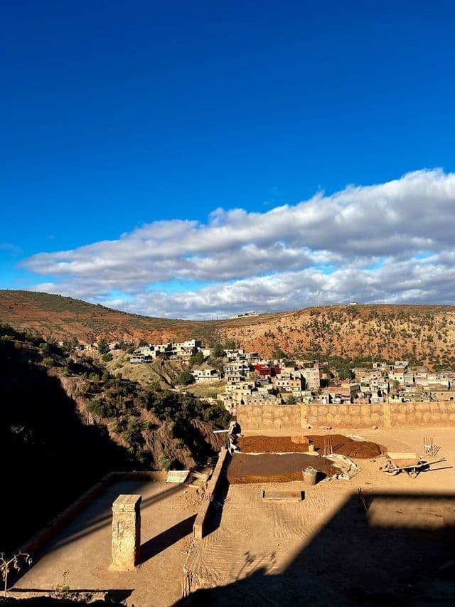 Un pueblo de edificios de color tierra anidado en colinas áridas y onduladas bajo un cielo azul brillante con nubes dispersas.