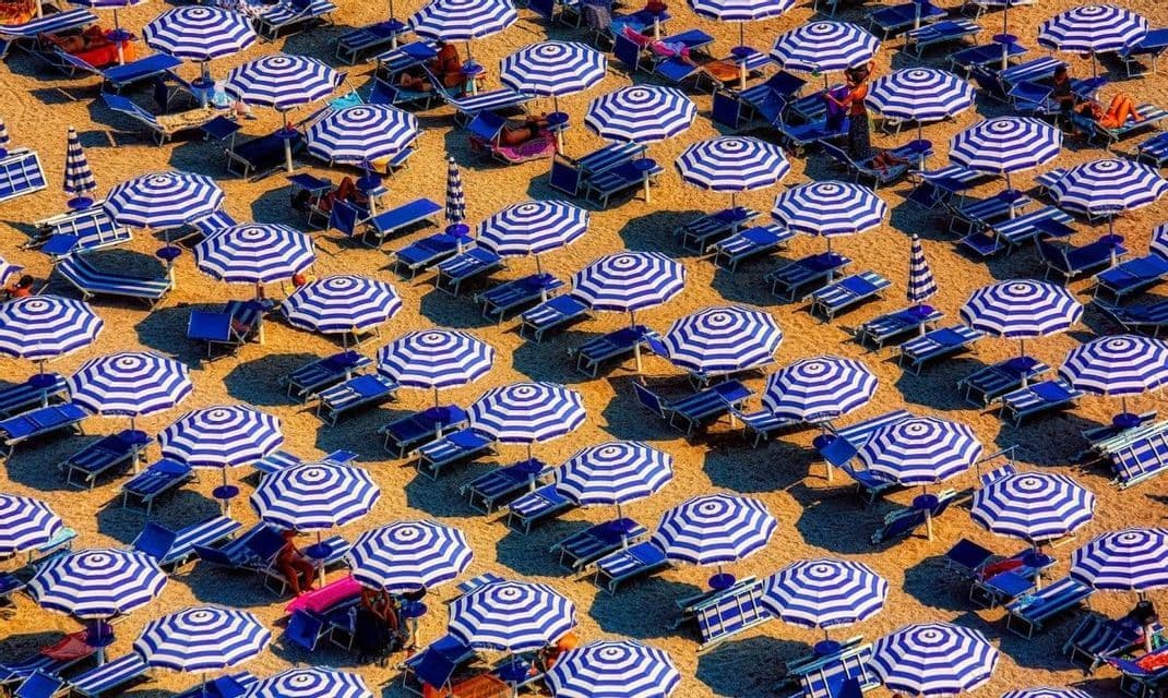 Una vista aerea di una spiaggia coperta da un motivo di ombrelloni a righe blu e bianche e lettini blu abbinati.