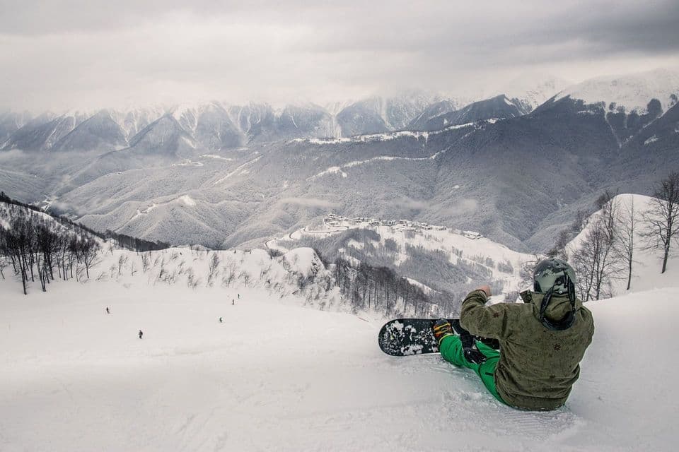 Uno snowboarder con il casco siede su un pendio innevato, osservando una vasta catena montuosa coperta di neve sotto un cielo nuvoloso.