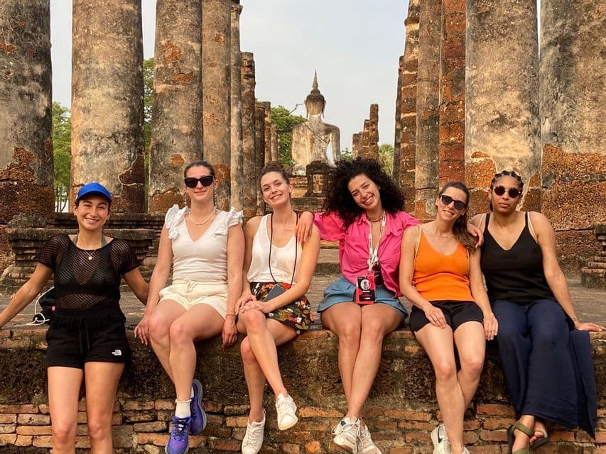Un viaje en grupo de WeRoad con seis mujeres sentadas en un muro de ladrillo, posando para una foto frente a antiguas ruinas de templo con una estatua de Buda.