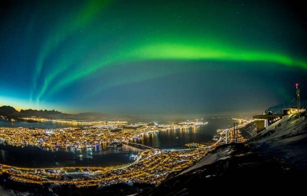 Las auroras boreales verdes se extienden por el cielo nocturno sobre una ciudad costera brillantemente iluminada rodeada de agua y montañas nevadas.