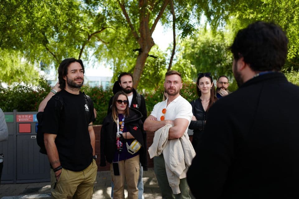 Un gruppo WeRoad in viaggio si trova all'aperto sotto alberi verdi, ascoltando attentamente un oratore in primo piano.