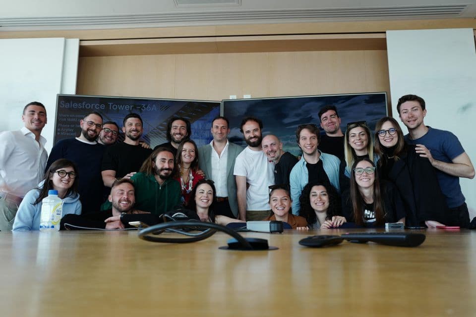 Un folto gruppo di persone sorridenti in posa per una foto di gruppo dietro un lungo tavolo da conferenza in un ufficio.
