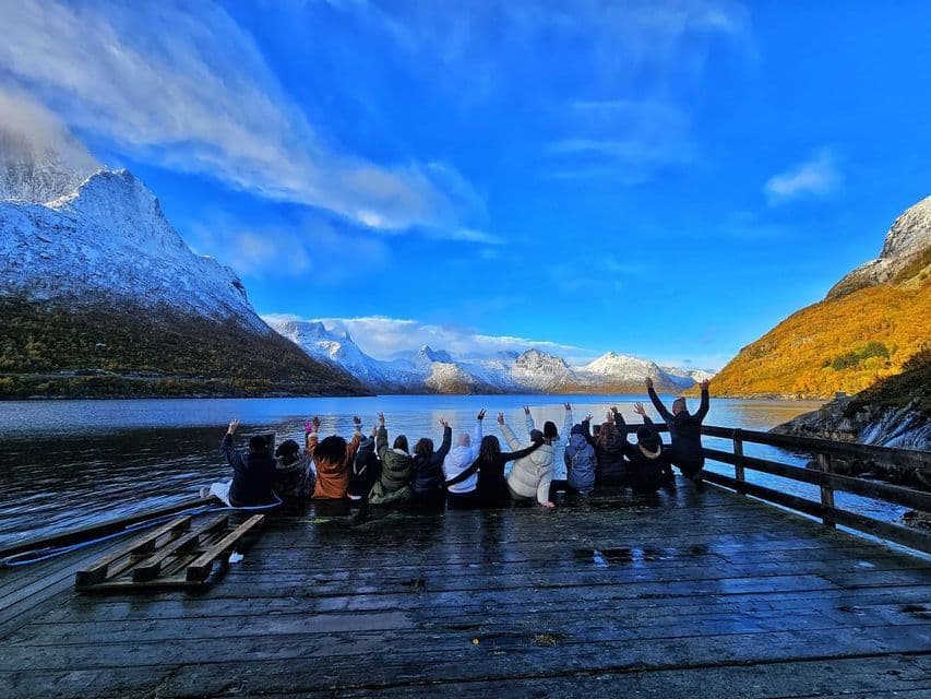 Un grupo de WeRoad, de viaje, se sienta en un muelle de madera mojado con los brazos en alto, frente a un fiordo y montañas nevadas bajo un cielo azul.