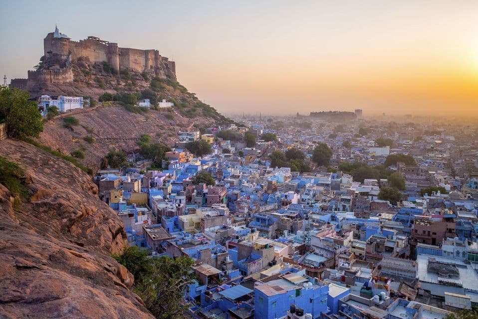 A large stone fort sits on a rocky hill, overlooking a dense city with many blue buildings at sunrise.