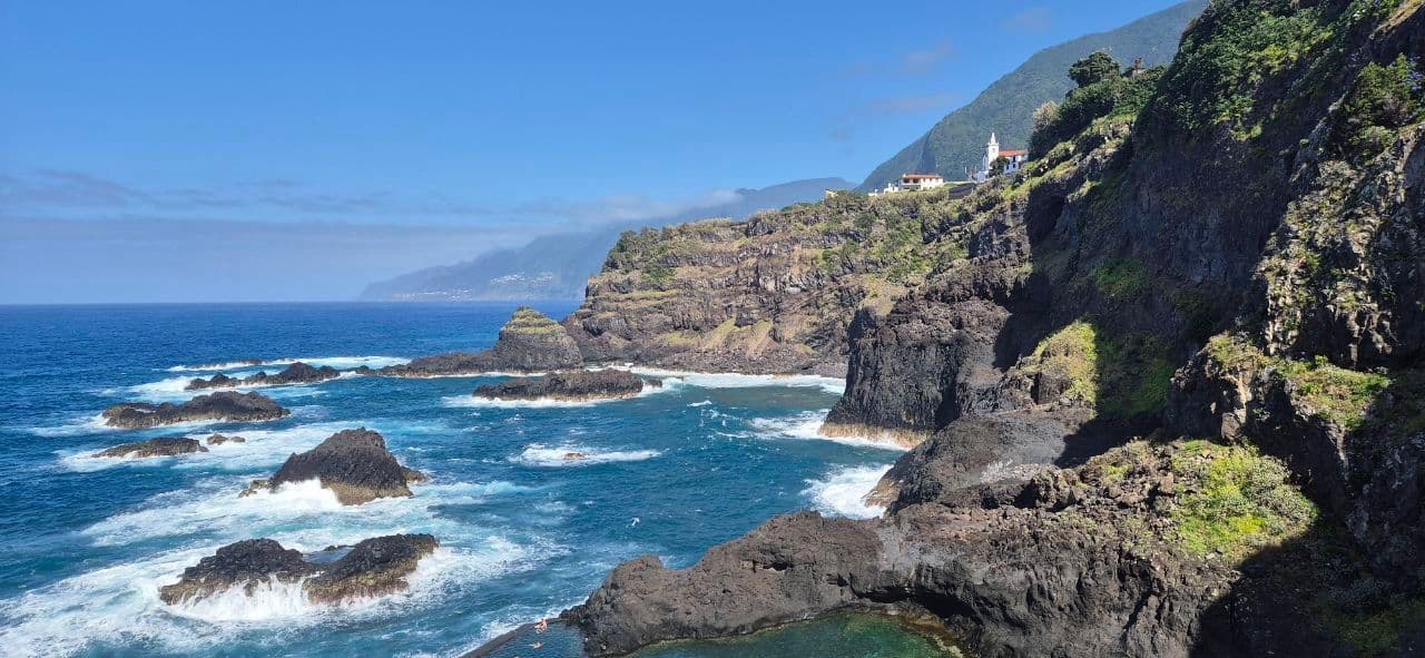 Onde si infrangono su una costa vulcanica aspra, con un piccolo villaggio e una chiesa visibili sulle verdi scogliere sotto un cielo azzurro e limpido.
