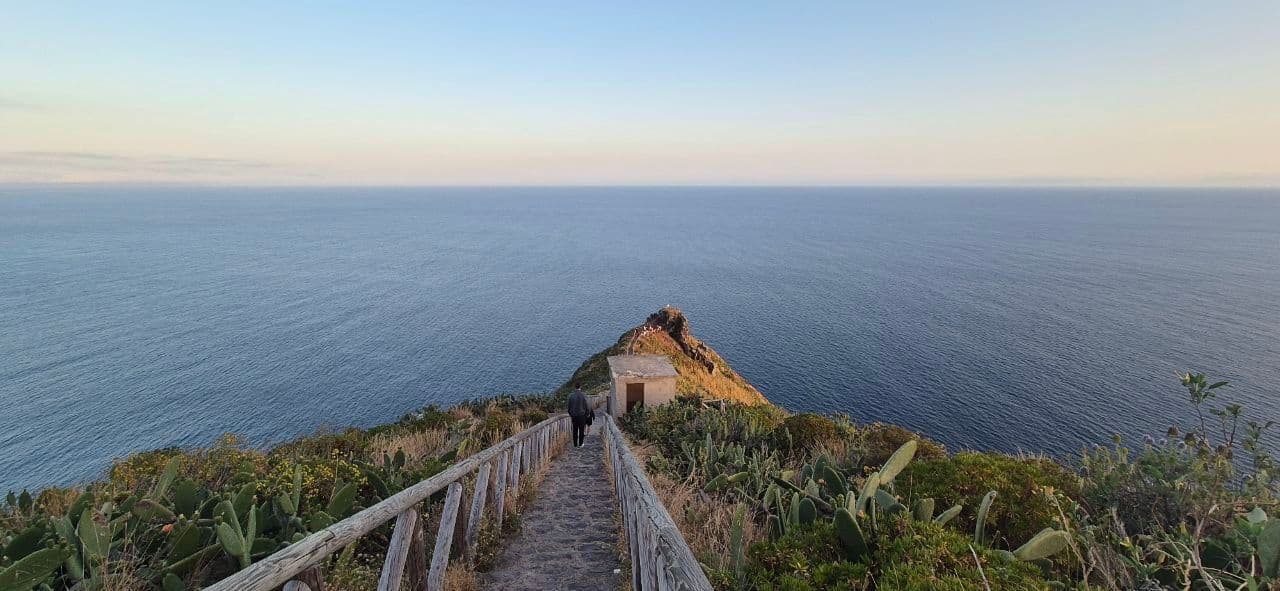 Una persona cammina lungo un sentiero in pietra con un parapetto in legno su un promontorio costiero che si affaccia su un vasto mare al tramonto.