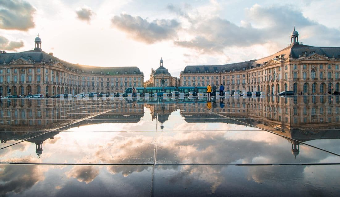 Un complejo de edificios históricos se refleja en la superficie mojada de una gran plaza bajo un cielo nublado al atardecer.