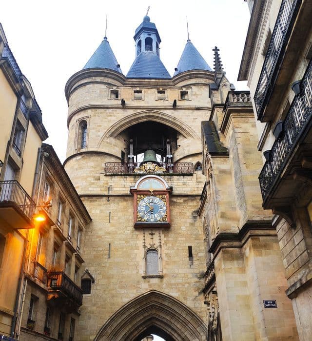 Una vista en contrapicado de una torre del reloj de piedra con dos torretas puntiagudas, una campana grande y una esfera de reloj ornamentada, vista desde una calle estrecha.