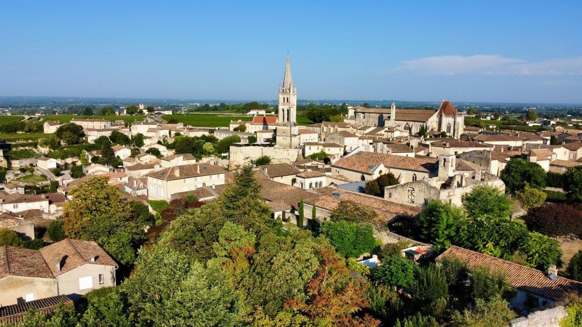 Una vista aérea de un pueblo europeo histórico con un prominente campanario de iglesia, rodeado de frondosos árboles y campo bajo un cielo despejado.