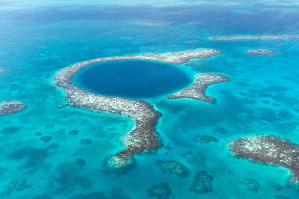 Vue aérienne d'un grand trou bleu profond et circulaire, entouré d'un atoll de récif corallien dans un océan turquoise.