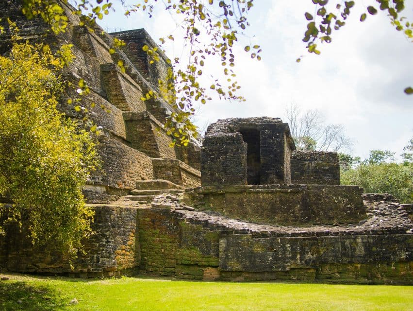 Une ancienne pyramide de pierre en ruine se dresse sur un champ herbeux, entourée d'arbres verdoyants, sous un ciel partiellement nuageux.