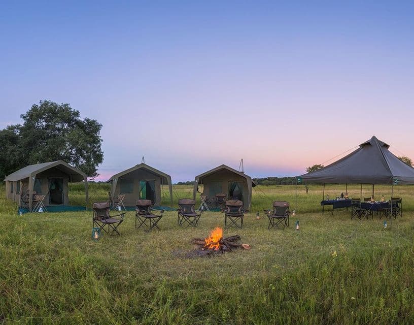Un campeggio con tende safari e sedie disposte intorno a un falò acceso in un campo erboso al tramonto.