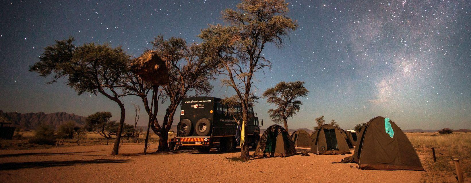 Un campeggio con un veicolo overland e tende montate in una savana sotto un cielo notturno stellato con la Via Lattea visibile.