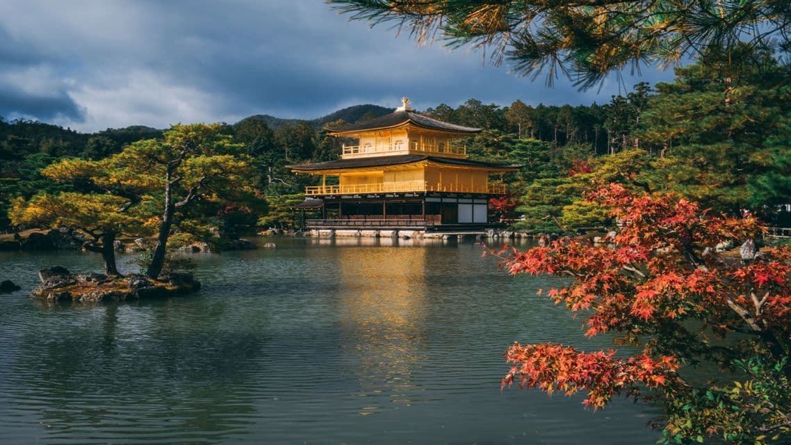 A golden Japanese temple sits beside a tranquil lake, surrounded by colorful autumn foliage and forested hills.