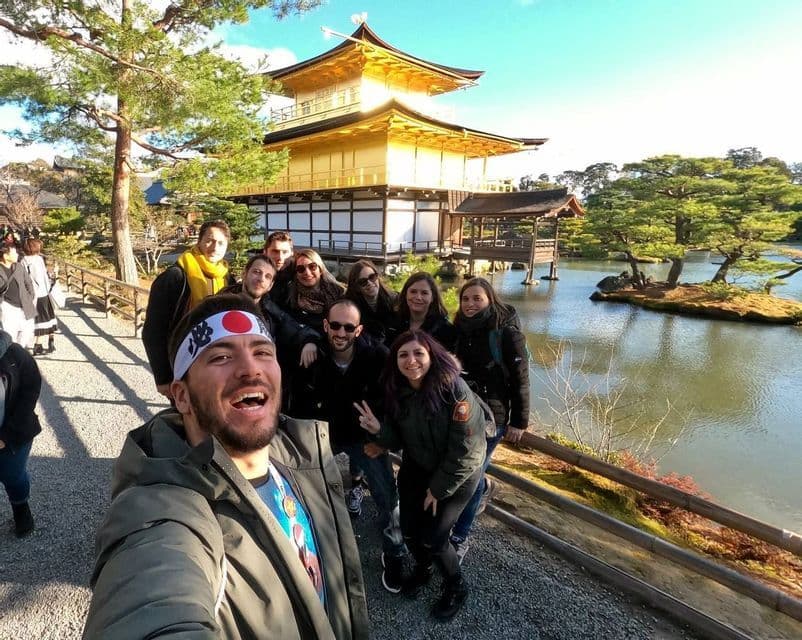 A WeRoad group trip takes a smiling selfie in front of a traditional golden temple next to a pond on a sunny day.