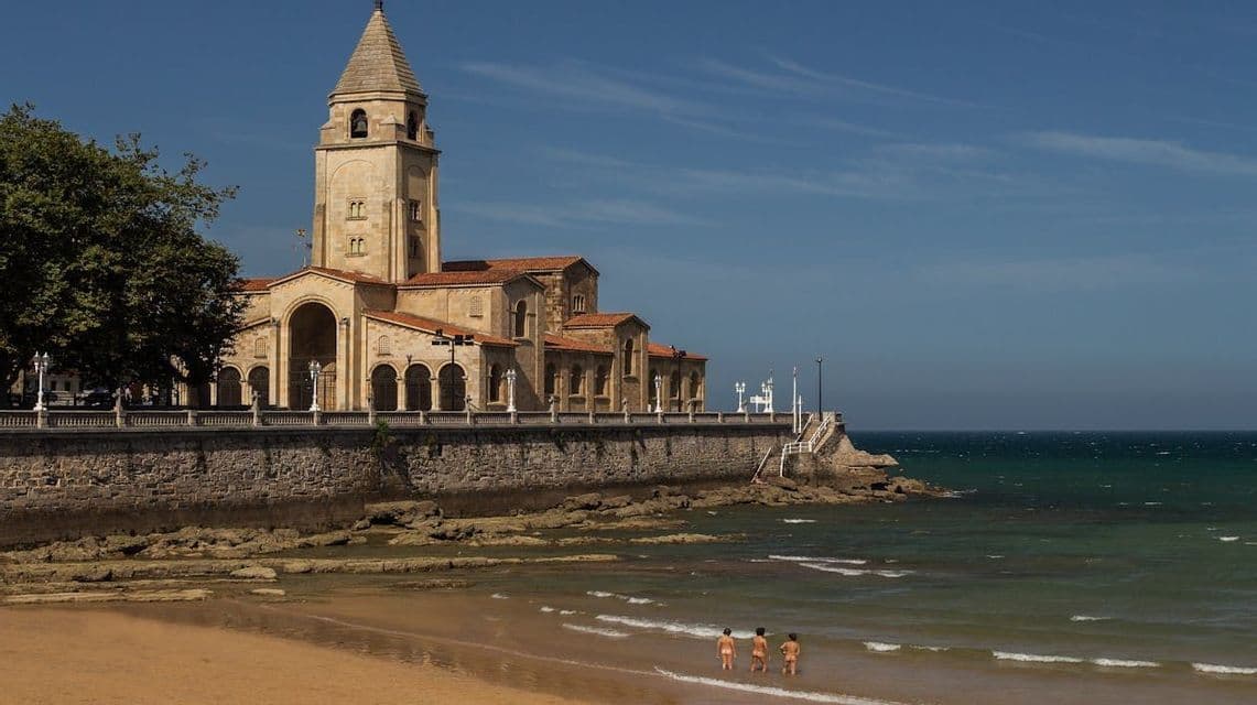 Una gran iglesia de piedra con un campanario se alza sobre un malecón que domina una playa donde tres personas vadean en el océano.