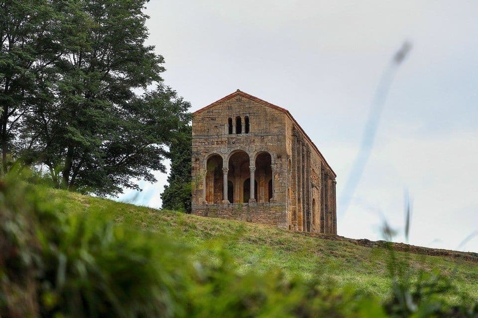 Un antiguo edificio de piedra con un pórtico de arcos abiertos se alza en una colina cubierta de hierba junto a un gran árbol bajo un cielo nublado.