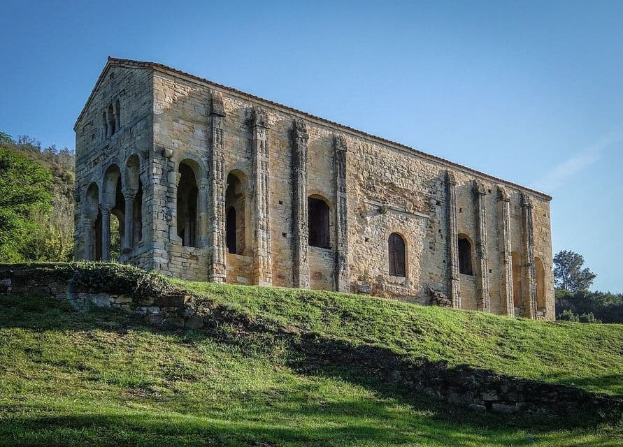 Un antiguo edificio de piedra con contrafuertes y ventanas arqueadas se asienta en una colina cubierta de hierba bajo un cielo azul claro.