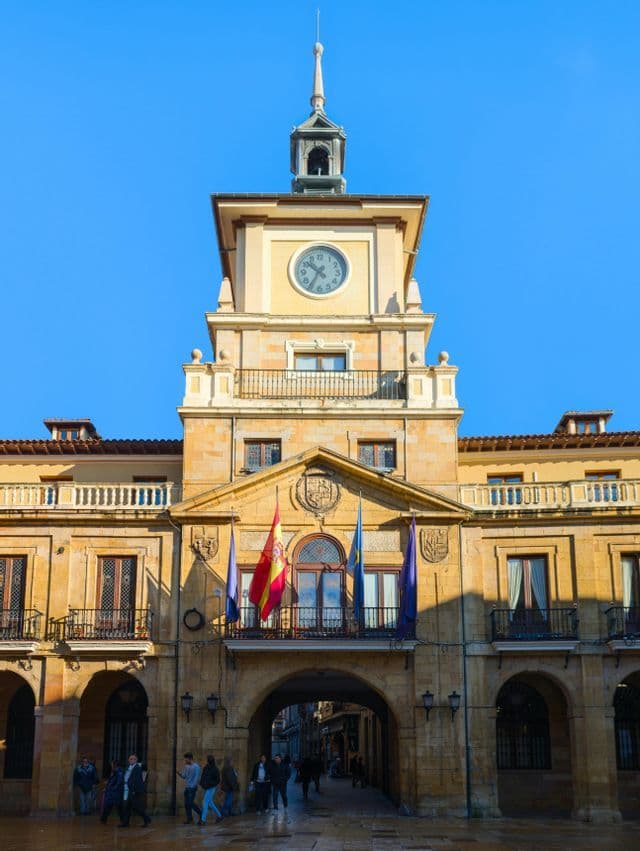 Una vista en contrapicado de un edificio municipal de piedra con una destacada torre del reloj y banderas ondeando desde un balcón, bajo un cielo azul despejado.