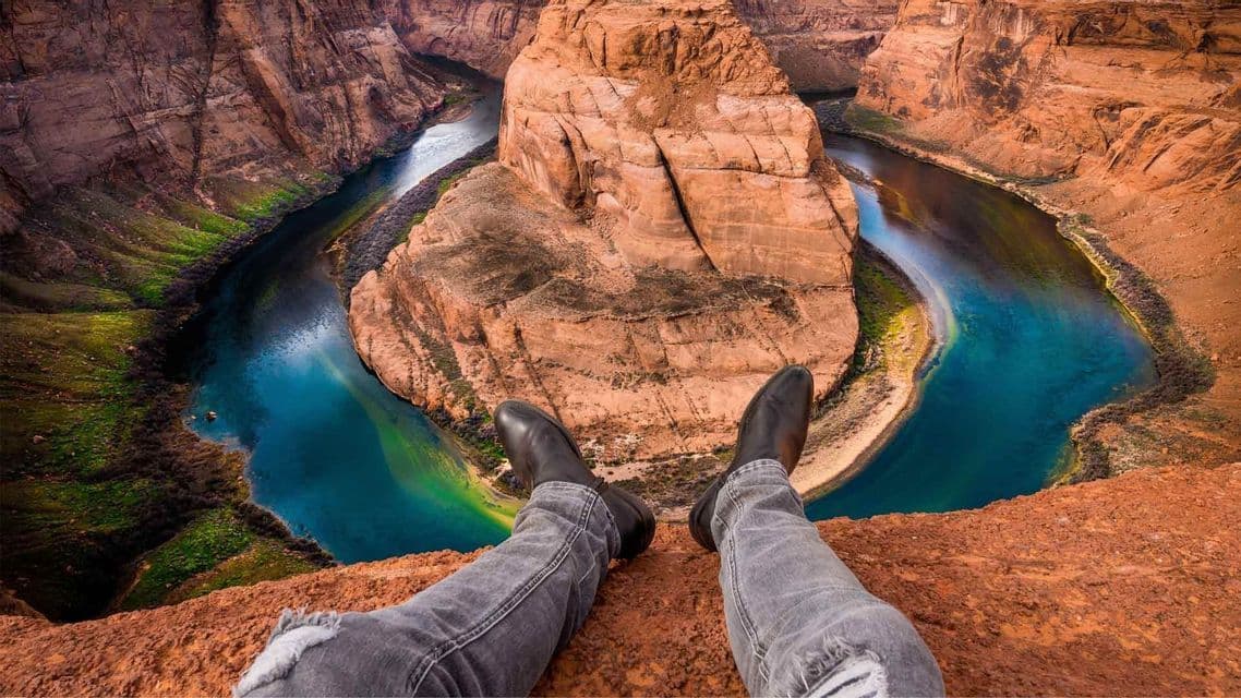 Gambe in jeans grigi e stivali neri, in prima persona, sul bordo di un canyon di roccia rossa con vista sull'ansa di un fiume.