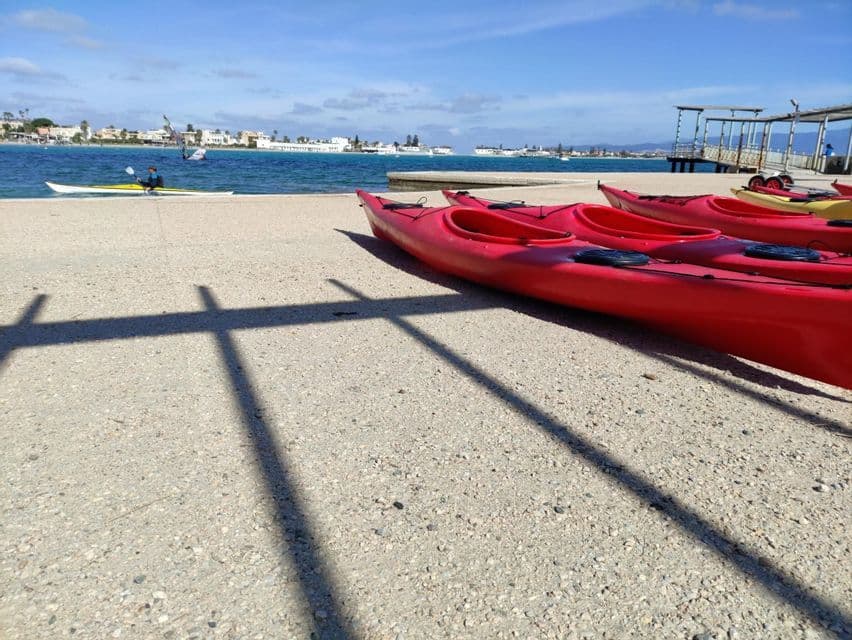 Varios kayaks rojos alineados en un muelle de hormigón junto al mar, con un pueblo al fondo.