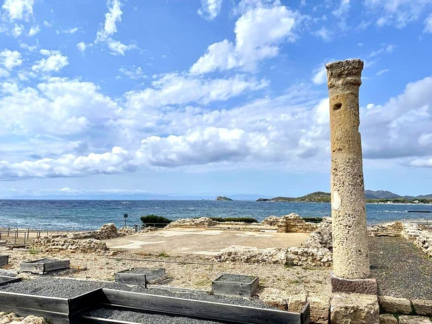 Una columna de piedra erosionada se alza entre ruinas arqueológicas junto al mar, bajo un cielo azul parcialmente nublado.