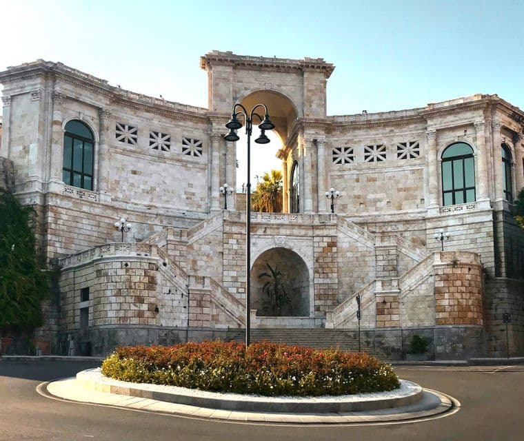Un gran monumento de piedra con un arco central y grandes escalinatas, visto desde una rotonda con un parterre de flores.