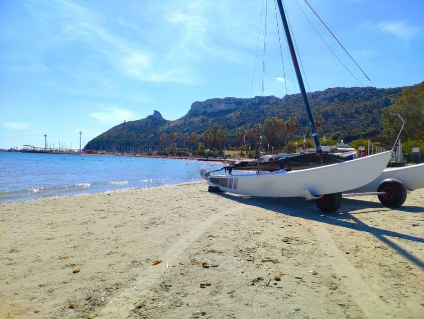 Un catamarán de vela blanco con ruedas se encuentra en una playa de arena, con una montaña verde y el mar de fondo.