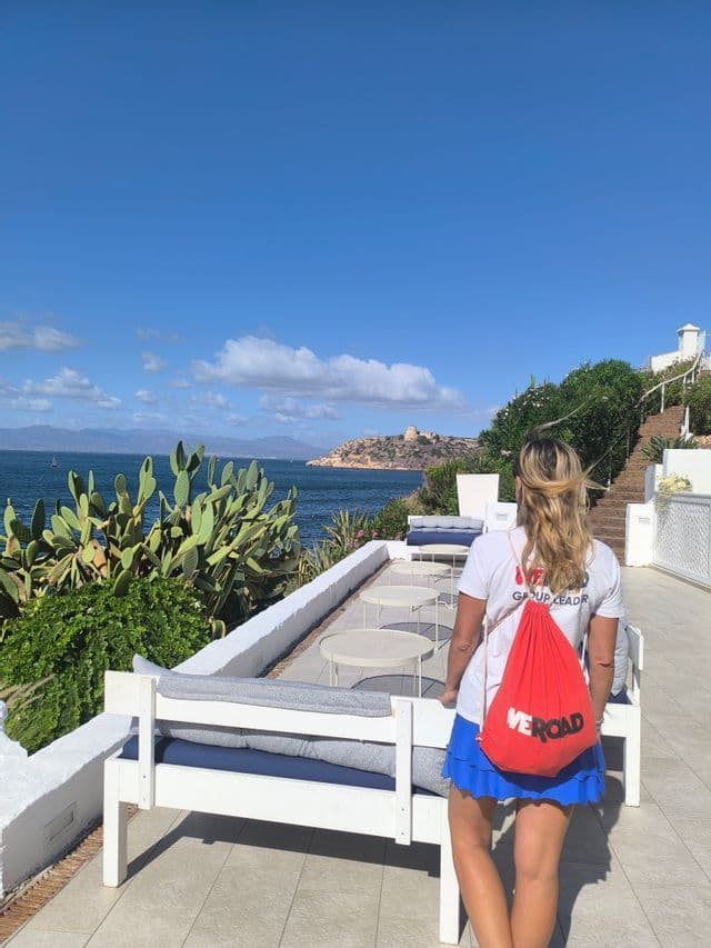 Un guía WeRoad visto de espaldas, de pie en una terraza con vistas al mar y un cabo distante en un día soleado.