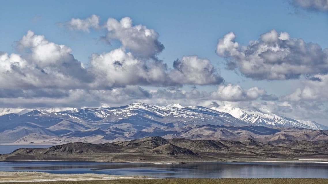 Un lago placido si adagia di fronte a colline brulle e a una lontana catena montuosa con cime innevate, sotto un cielo parzialmente nuvoloso.